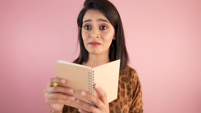 Close up portrait of shocked amazed young Asian woman student girl hold notebook diary reading learn feels astonishment shock look at camera, isolated over light pink studio background.
