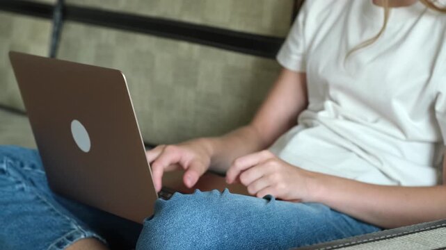 Teenager girl sitting on sofa and typing on laptop keyboard in casual clothes. Concept of online homework, remote work, blogging and social media.