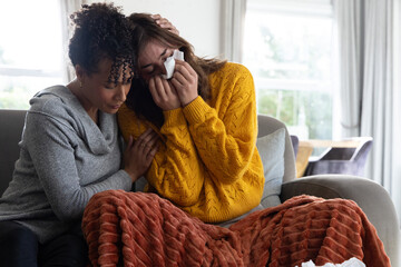 Fototapeta premium Diverse female friends sitting on gray couch in living room, comforting woman with tissue
