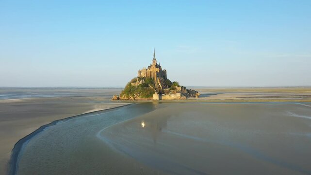 Aerial view of Mont Saint Michel island surrounded by tidal flats at low tide under a clear blue sky in Normandy, France.