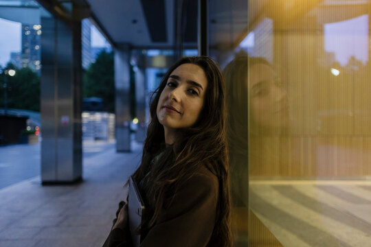Businesswoman leaning against office building with laptop at night