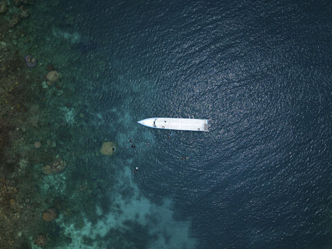 Aerial view of a lone white yacht glides on the dark blue sea near the vibrant greens and browns of the shore, Karimunjawa, Central Java, Indonesia.