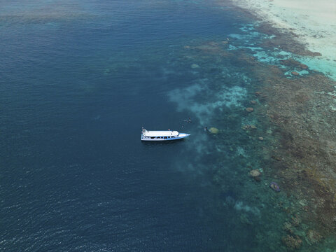 Aerial view of a lone white boat sailing on the deep blue sea near the colorful coral reefs of Cemara Kecil, Karimunjawa, Central Java, Indonesia.