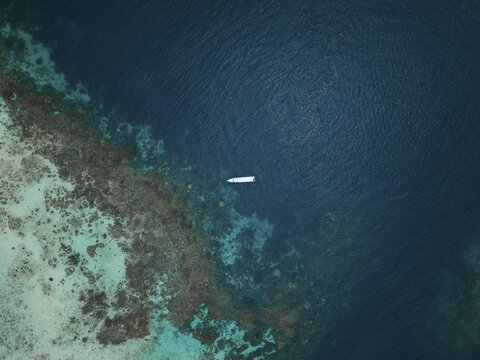 Aerial view of a boat floating on the contrasting dark blue sea alongside the lighter turquoise shore of Cemara Kecil Snorkeling Spot, Karimunjawa, Central Java, Indonesia.
