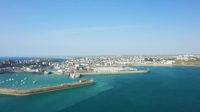 Scenic aerial view of Granville harbor with boats, marina, and coastal town under a clear blue sky. The turquoise sea and breakwaters frame the picturesque French shoreline.