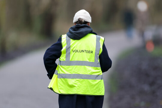 A volunteer viewed from behind wearing a bright yellow high-vis vest marked "VOLUNTEER," standing on a wet tarmac park path with a softly blurred bokeh background of bare winter trees and parkland