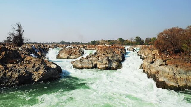 Wide view of Khone Phapheng Falls with powerful rapids rushing through rocky terrain under a clear sky in Laos. Dramatic natural landscape with vibrant water and rugged rocks.