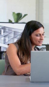 Vertical video: Reacting to peer, woman in brown top reviewing plan with laptop, 3D plan in studio