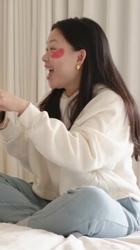 Vertical video: Speaking woman in white hoodie touching red patch on bed, holding glass, offering