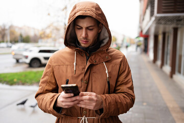 A young man wearing a brown hooded jacket stands on an urban sidewalk and looks at his smartphone screen during the day. © Dusko
