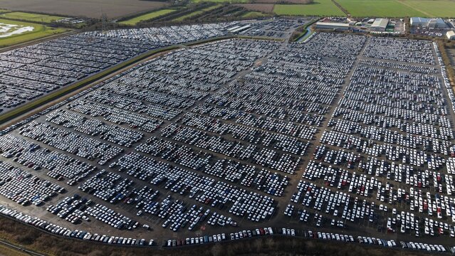 Aerial view of a vast grid of neatly arranged vehicles stretching towards the horizon, punctuated by roads and industrial structures, Grimsby, United Kingdom.