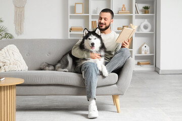 Handsome young man sitting on sofa and reading book with cute husky dog in living room © Pixel-Shot