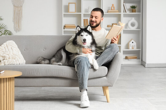 Handsome young man sitting on sofa and reading book with cute husky dog in living room