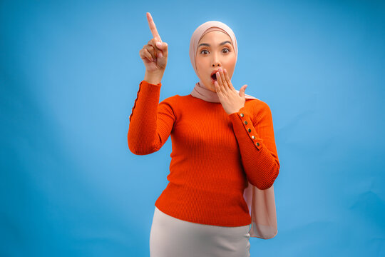 Surprised young Asian Muslim woman in hijab pointing to the copy space upwards.