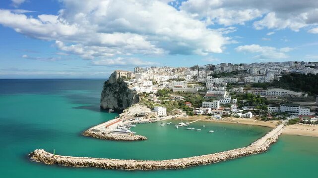 Scenic aerial view of the coastal town of Peschici, Italy, featuring turquoise waters, a marina, and whitewashed buildings on a cliff under a partly cloudy sky.