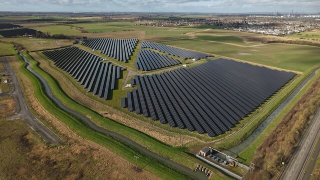 Aerial view of gleaming solar panels arranged in precise rows against a backdrop of green fields and distant buildings, Immingham, United Kingdom.