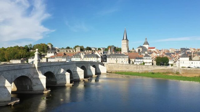 Scenic view of the historic stone bridge and charming old town of La Charite sur Loire, France, under a clear blue sky. The tranquil river and traditional architecture create a picturesque landscape.