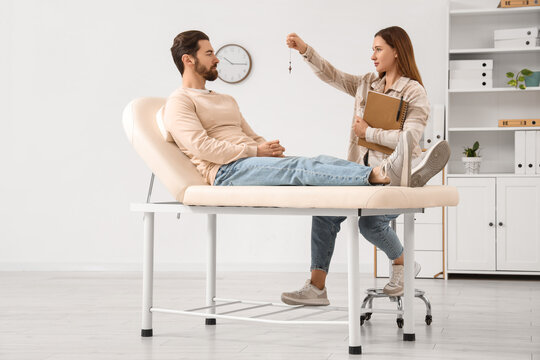 Female psychologist with pendulum hypnotizing young man on couch in office