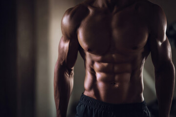 Obraz premium faceless muscular man facing camera showing defined abs, strong torso, neutral pose, minimal gym background softly blurred, dramatic side lighting highlighting core muscles, shallow depth of field.