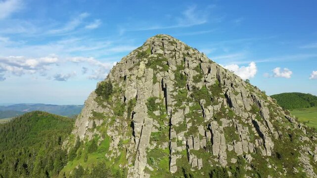 Aerial view of the rugged rocky summit of Mont Gerbier de Jonc in Ardeche, France, under a clear blue sky. Lush green vegetation surrounds the dramatic volcanic formation.