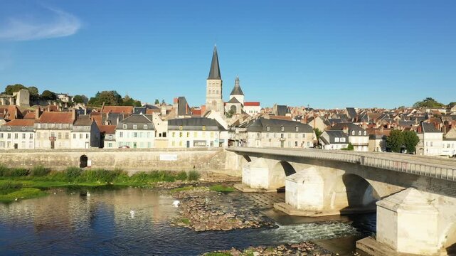 Scenic view of La Charite sur Loire with its iconic stone bridge, traditional houses, and church under a clear blue sky. The Loire River flows gently beneath the historic architecture.