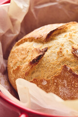 Golden sourdough bread with cracked crust in red ceramic Dutch oven, lined with parchment paper. Perfect for culinary, baking, and food photography projects.
