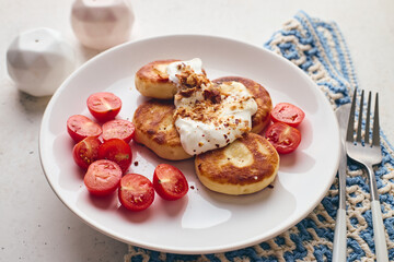 Savory syrniki (cheese pancakes) topped with sour cream, crispy fried onions, and fresh cherry tomatoes, served on a white plate with a blue knit cloth. Perfect for breakfast or brunch.