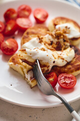 Savory syrniki (cheese pancakes) topped with sour cream, crispy fried onions, and fresh cherry tomatoes, served on a white plate with a blue knit cloth. Perfect for breakfast or brunch.