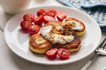 Savory syrniki (cheese pancakes) topped with sour cream, crispy fried onions, and fresh cherry tomatoes, served on a white plate with a blue knit cloth. Perfect for breakfast or brunch.
