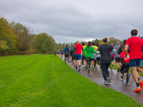 A large group of runners viewed from behind running along a glistening wet tarmac park path on an overcast autumn morning, with vivid green grass verges and golden autumn woodland lining the route