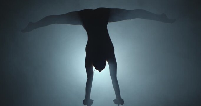 Full shot silhouette of a young circus gymnast performs a arms handstand on canes in a dark studio against a spotlight. For acrobatics, circus show and video production. Slow motion.