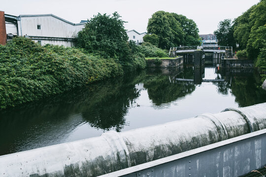 Calm canal water reflection beside trees and greenery with industrial warehouse on Hamburg Germany riverbank in soft daylight