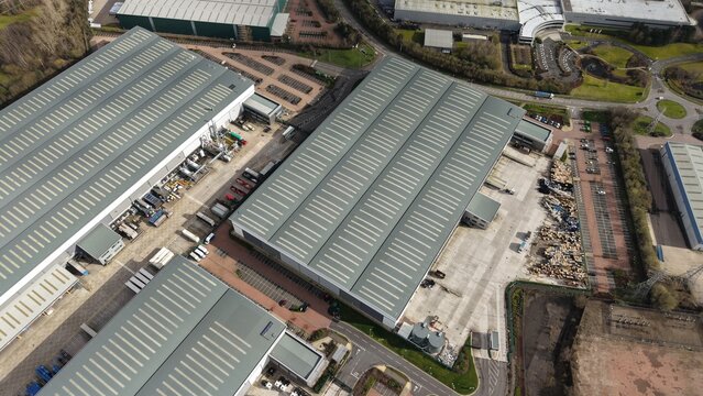 Aerial view of industrial buildings casting long shadows under a pale sky, amidst neatly organised parking and bordered by verdant trees, Birmingham, England, United Kingdom.