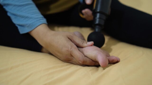 Anonymous woman massaging her foot with a portable electric massage gun at home. Close-up of female foot receiving percussion therapy for pain relief and muscle recovery