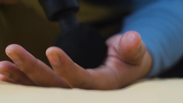 Anonymous woman massaging her palm and hand using a portable electric massage gun at home. Close-up of female hand receiving percussion therapy for muscle tension relief and recovery