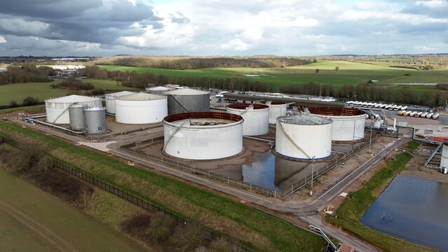 Aerial view of gleaming white and silver tanks contrast sharply with the surrounding green fields and dark, brooding sky at Kingsbury Oil Terminal, Kingsbury, Tamworth, United Kingdom.
