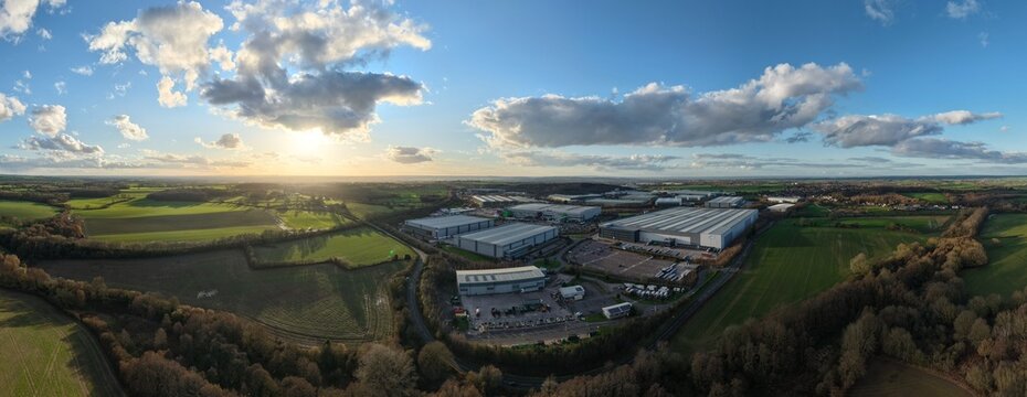 Aerial view of golden sunlight bathes verdant fields contrasting with the sharp lines of industrial buildings under a vast, cloud-strewn sky, Tamworth, England, United Kingdom.
