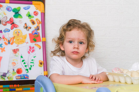 Moscow, Russia, 12.04.2025 A young child sits at a table with a white shirt on. The child is looking at the camera with a sad expression. There are several toys and a butterfly sticker on the wall beh