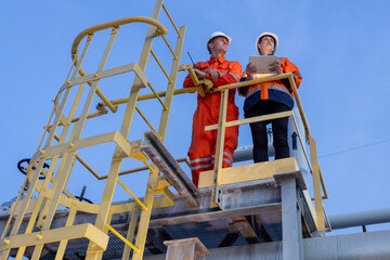 group of industrial engineers workers in a refinery - oil and gas processing equipment and machinery, engineers collaborate with a laptop, blueprint, and digital tablet at the oil storage tanks site.