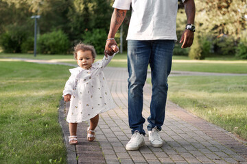 Fototapeta premium Father holding little daughter hand while child takes first steps on park path. Dad with baby outdoors