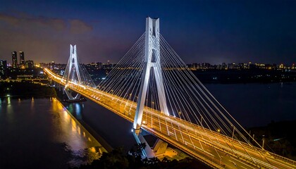 Obraz premium Illuminated bridge spans water at night, connecting city skyline in the distance with blurred streaks of traffic
