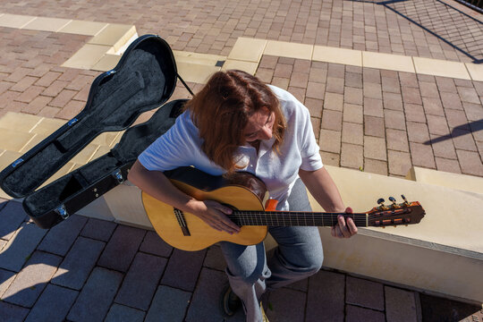 Woman playing acoustic guitar outdoors, busking performance with open case on brick pavement