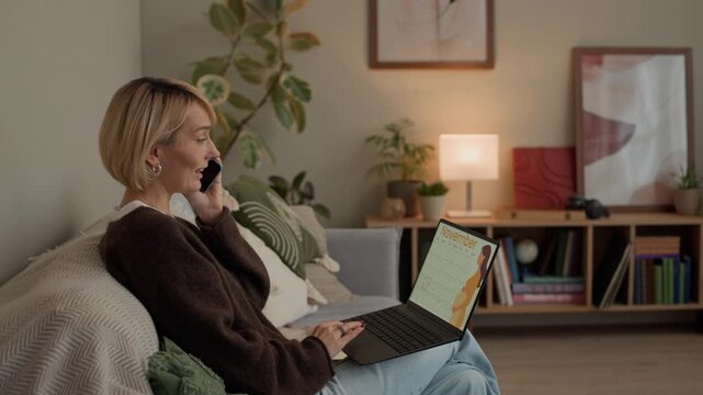 Handheld shot of adult Caucasian woman sitting on couch calling on smartphone and checking pregnancy calendar on wireless laptop in cozy living room