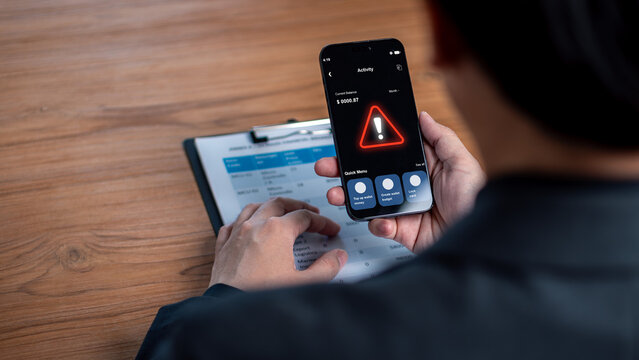 Cybersecurity risk and financial banking alert concept. Businessman holding a smartphone showing a red warning triangle icon over business documents.