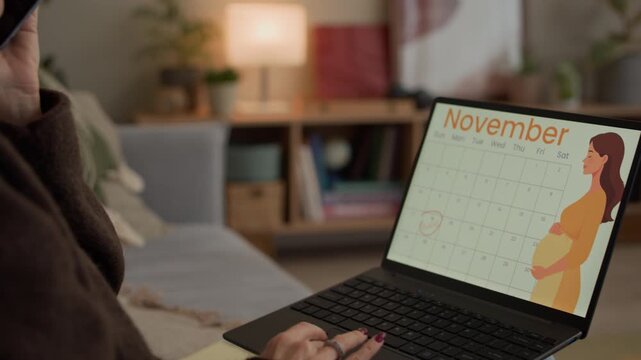 Tilt down shot of cheerful middle-aged Caucasian woman communicating on mobile phone while using wireless laptop with pregnancy tracker application in living room