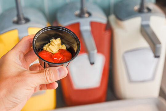 Hand holding a small plastic cup filled with red ketchup and yellow mustard, with condiment dispensers in the background