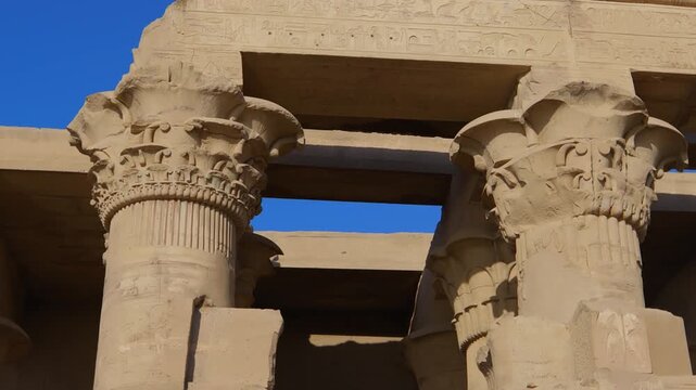 Kom Ombo temple columns with composite capitals and hieroglyphic carvings viewed from below against blue sky in Egypt, upward perspective of ancient sandstone architecture.