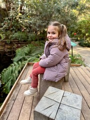 A teenage girl sitting on a park bench, looking directly at the camera, soft natural daylight, relaxed and natural pose, background of green trees and flowers, atmospheric and realistic outdoor photo