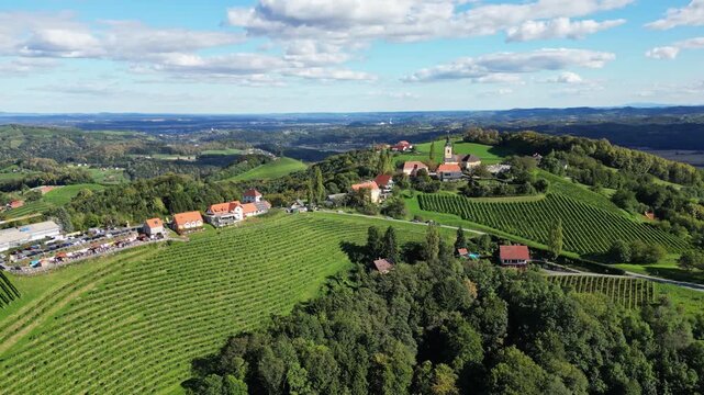Aerial view of Kitzeck im Sausal village nestled among green vineyards and rolling hills under a blue sky with white clouds, Kitzeck im Sausal, Styria, Austria.