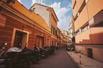 Colorful buildings line a narrow street where numerous scooters are parked. Fluffy clouds drift above, creating a lively atmosphere in this charming neighborhood © Cristina